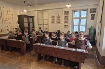 Učenci v starinskih oblekah sedijo v šolskih klopeh v Šolskem muzeju Slovenije